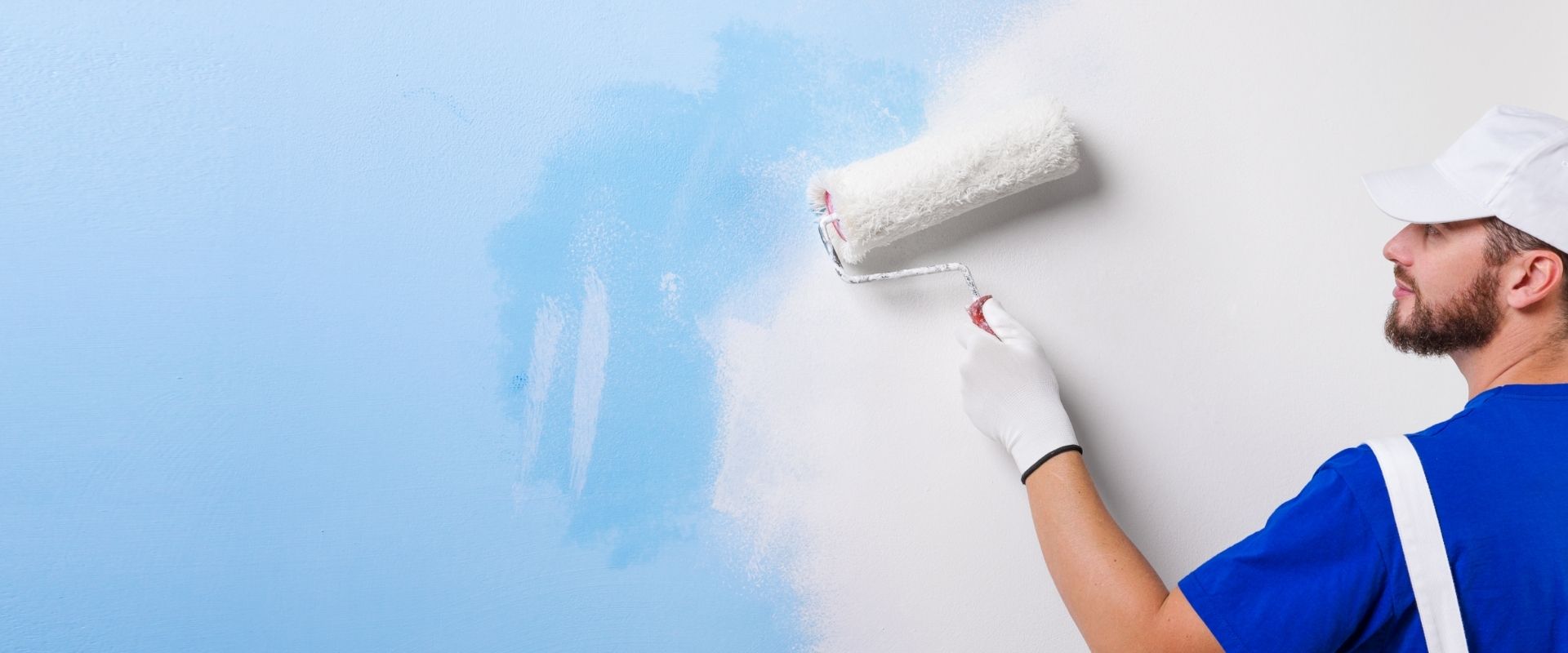 Eine Person in einem blauen Hemd und weißen Handschuhen streicht eine Wand mit einer Rolle und trägt dabei eine hellblaue Farbe auf.